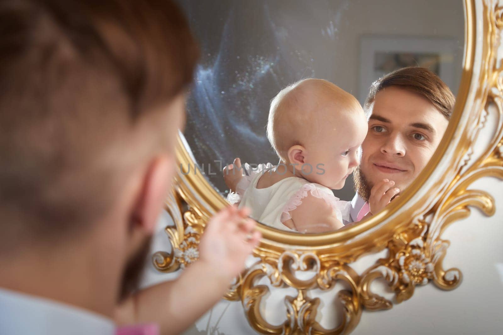 A father smiles while holding his baby daughter, who plays with her fingers, in a warm, inviting space.