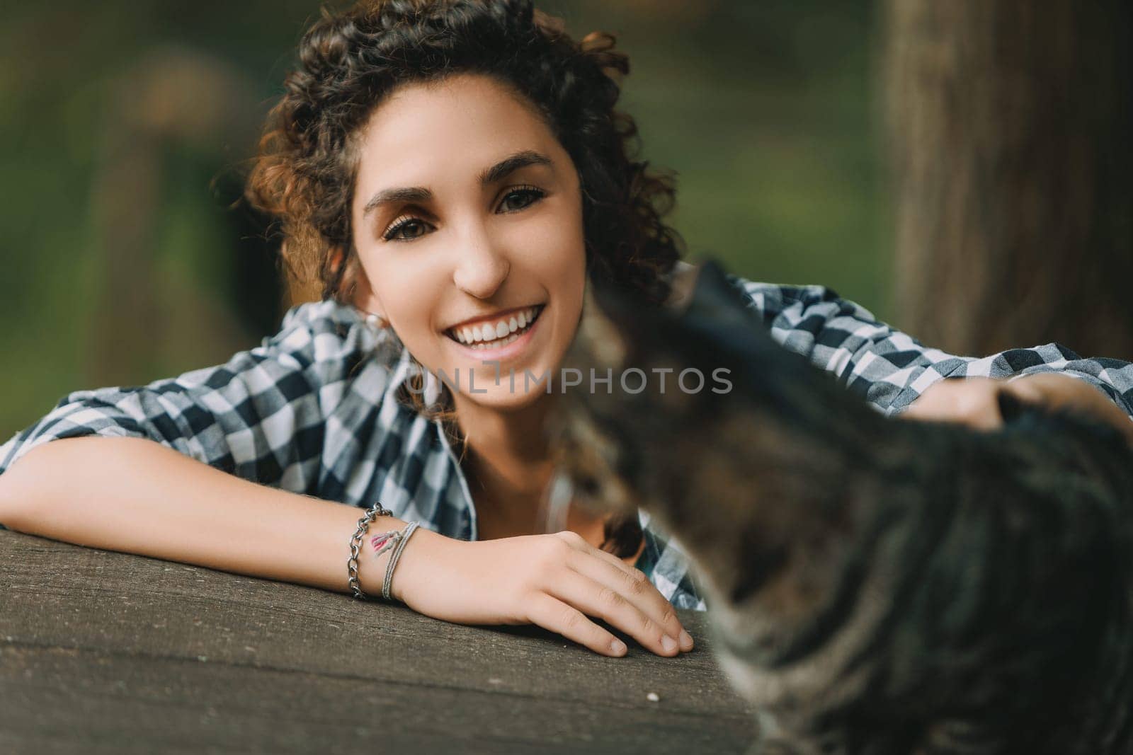 Curly-haired woman in a checkered shirt extends her arm to a tabby cat, smiling brightly. The cat appears slightly out of focus, emphasizing the woman's vivid, happy expression