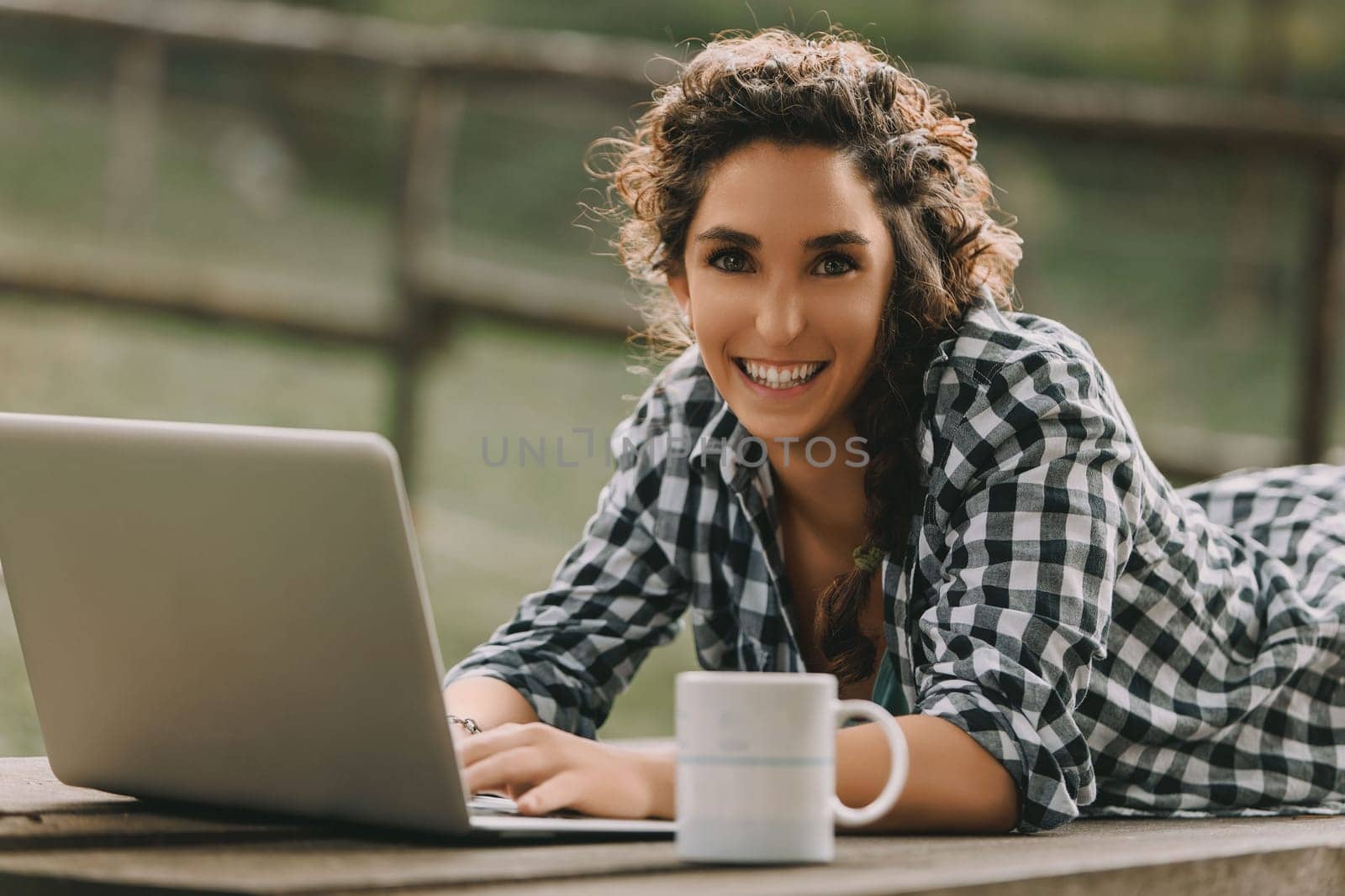 Woman with curly braided hair and a checkered shirt lies on her stomach, smiling at the camera while using a laptop. A coffee mug nearby adds to the casual, outdoor workspace vibe