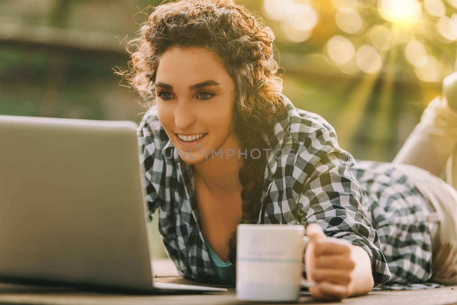 Curly-haired woman with a braid and checkered shirt lies on her stomach, typing on a laptop. Her cheerful smile and coffee mug convey a pleasant and engaging work environment