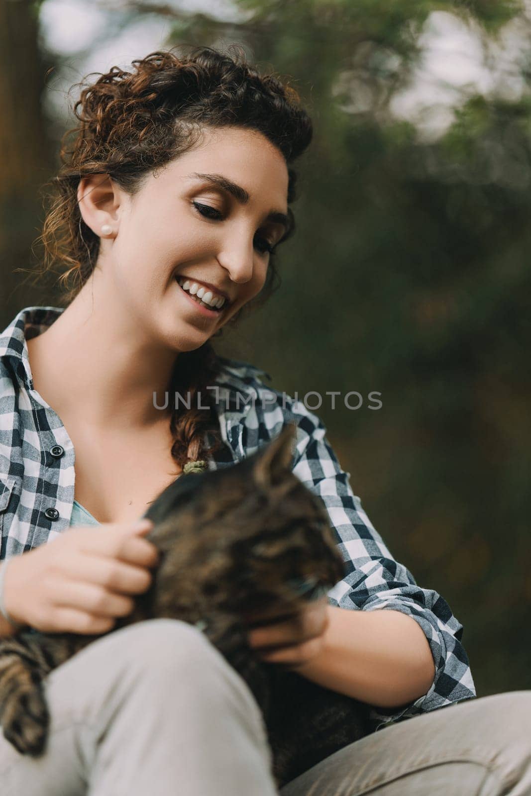young woman in a checkered shirt with curly hair holds a content tabby cat, smiling brightly. The cat’s expression is one of utmost relaxation, as if in a spa for cats