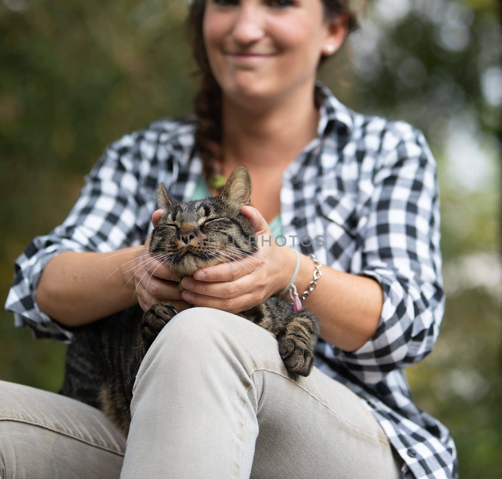 Woman with curly hair and checkered shirt smiles while holding a tabby cat. The cat looks incredibly relaxed, enjoying the moment as if receiving a luxurious spa treatment