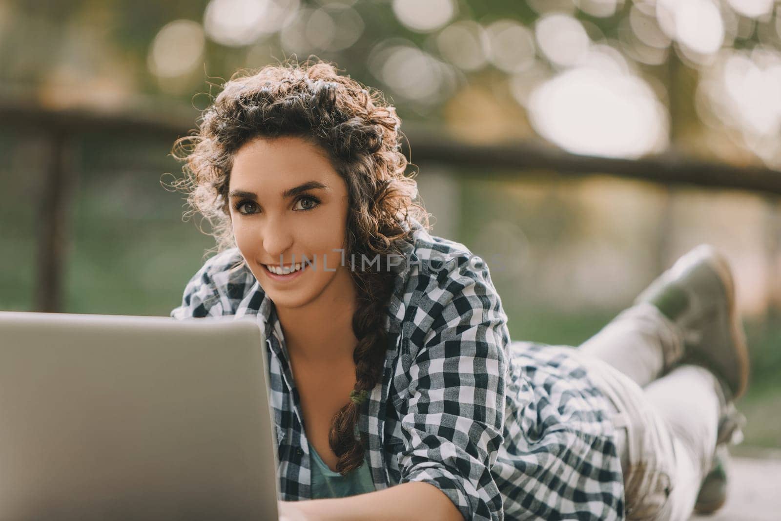 Woman with braided curly hair, wearing a checkered shirt, uses a laptop while lying on her stomach outdoors. Her concentrated expression and the warm sunlight create a serene working environment