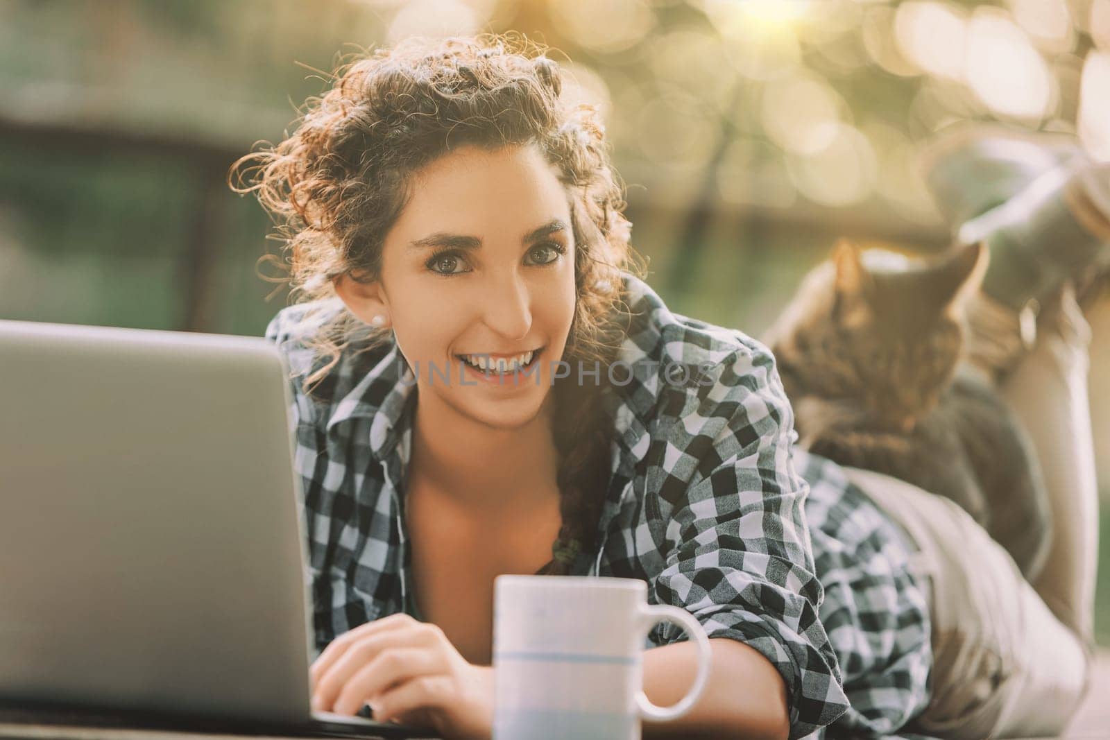 woman with curly braided hair and a checkered shirt lies on a bench, typing on a laptop. Her cat rests on her back, and a coffee mug is beside her, adding to the cozy scene