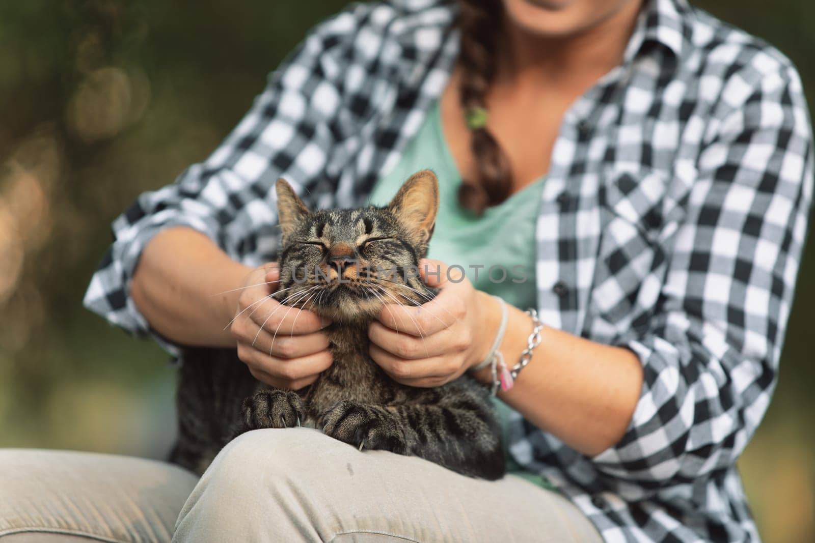 woman in a checkered shirt smiles, holding a delighted tabby cat. The cat’s relaxed demeanor suggests it is enjoying a spa-like pampering session