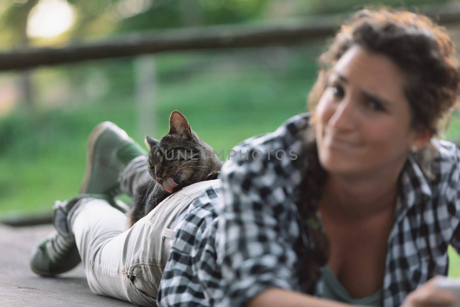 Young woman with curly hair in a braid lies down, her face out of focus, while a cat on her back grooms itself. The cat's relaxed demeanor indicates it feels very safe and secure