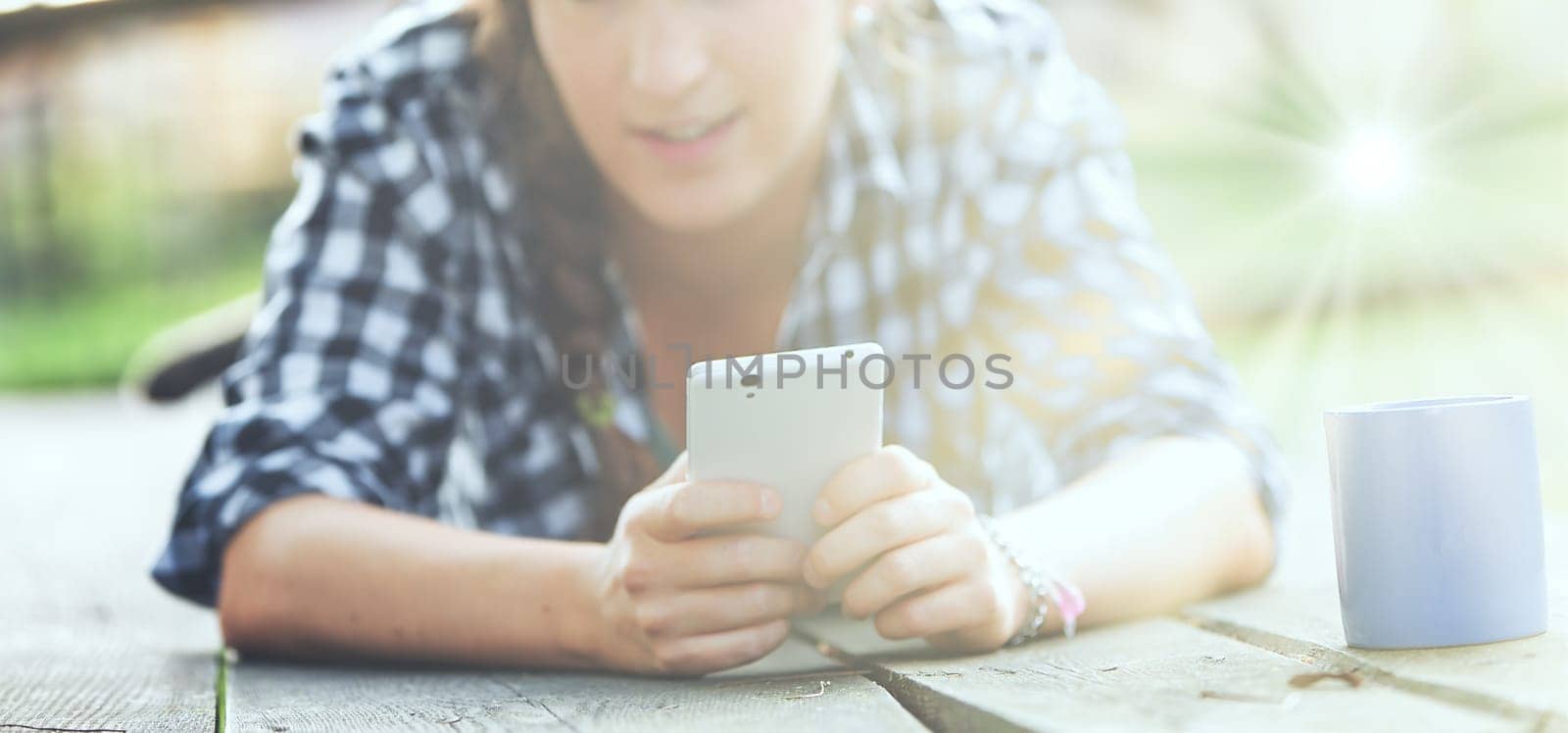 Woman with curly braided hair and a checkered shirt lies on her stomach outdoors, focusing on a smartphone. Her unrecognizable face and a blue mug create a peaceful, private scene