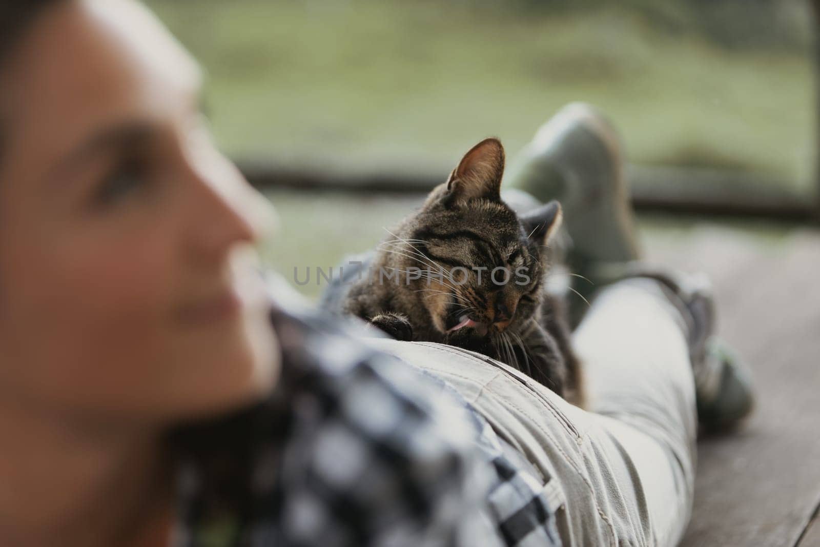 Woman with curly braided hair lies outdoors, her face blurred while a cat comfortably rests on her back, grooming itself. The cat's relaxed behavior reflects its sense of safety and trust