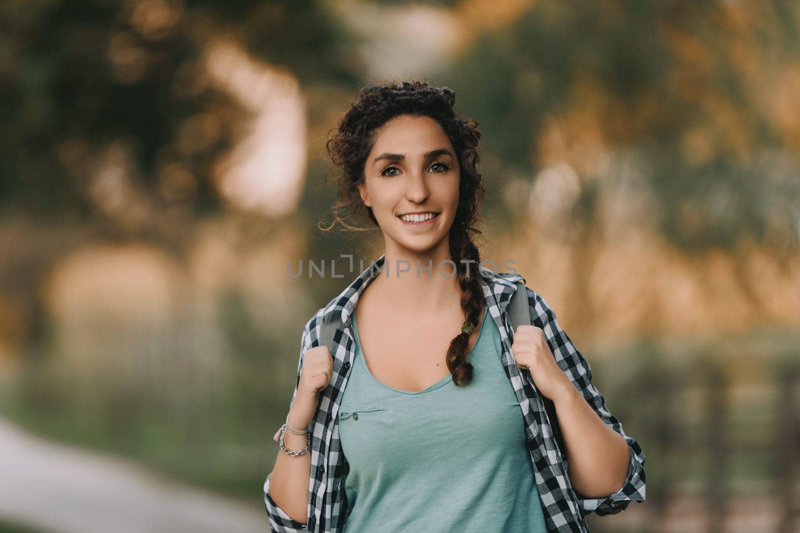 Young woman with curly braided hair and a checkered shirt smiles brightly while walking outdoors. Her joyful expression and relaxed posture highlight a serene and pleasant outdoor setting