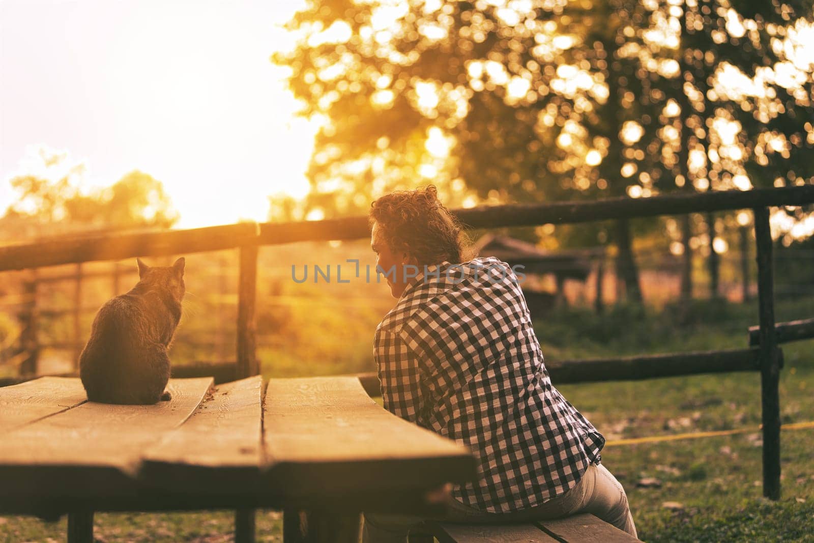 Woman with curly braided hair and a checkered shirt sits on a bench with her cat, both enjoying a serene sunset. Their relaxed postures reflect a peaceful retirement in a natural setting