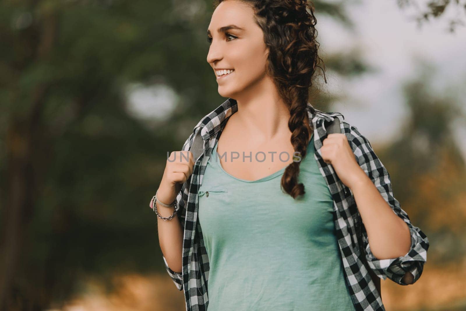 Young woman with braided curly hair, wearing a checkered shirt and green top, smiles brightly while walking outside. Her cheerful expression reflects a serene and pleasant environment