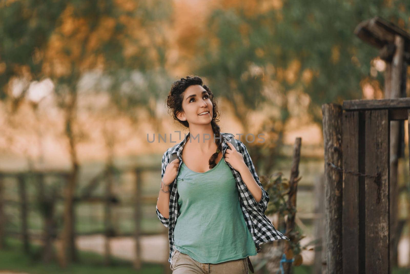 Curly-haired woman with a braid and checkered shirt smiles brightly while walking outside. Her cheerful demeanor and relaxed stance highlight a serene and pleasant environment