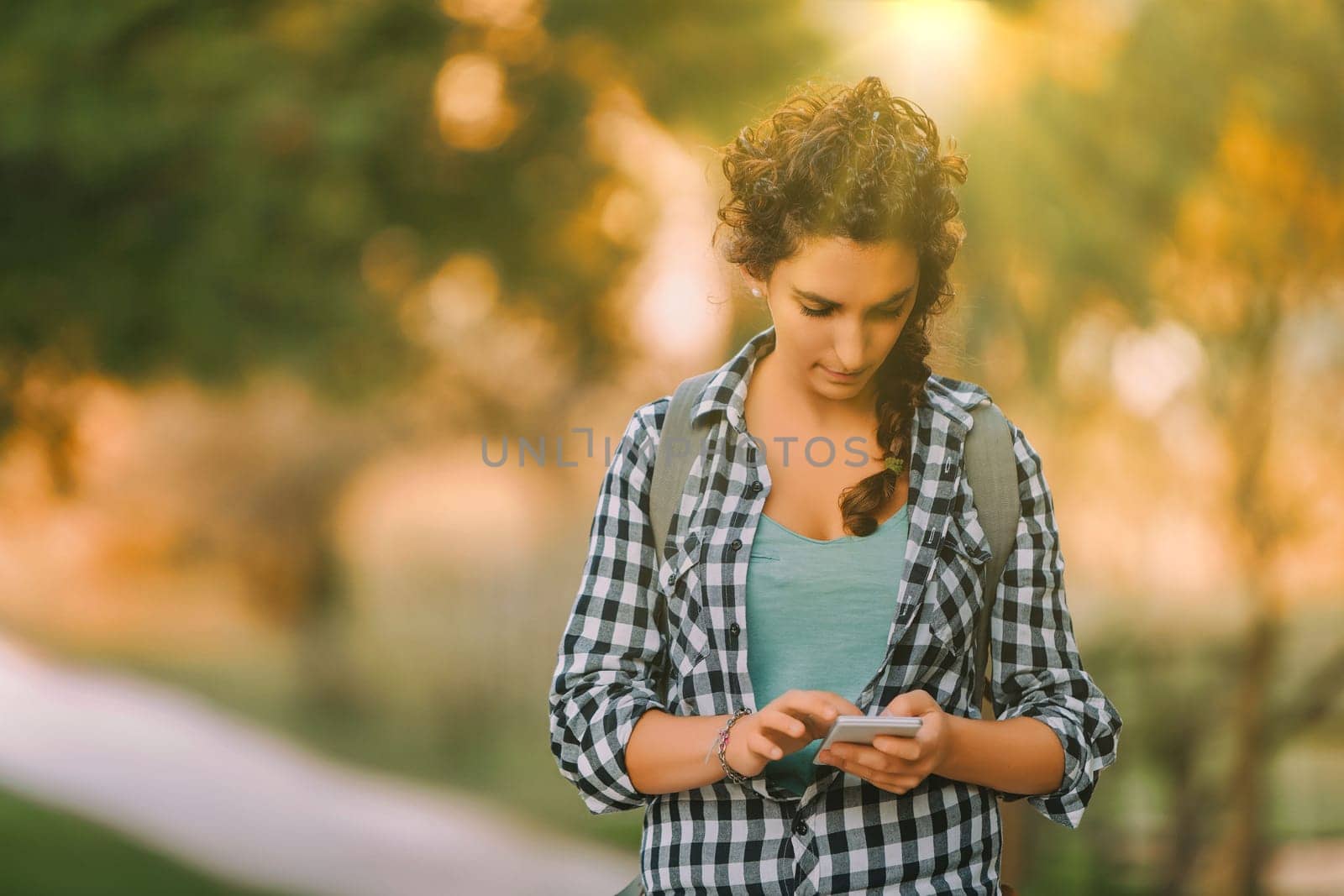 Woman with curly braided hair and a checkered shirt looks down at her smartphone while walking outdoors. Her focused expression and casual attire highlight her engagement with the device
