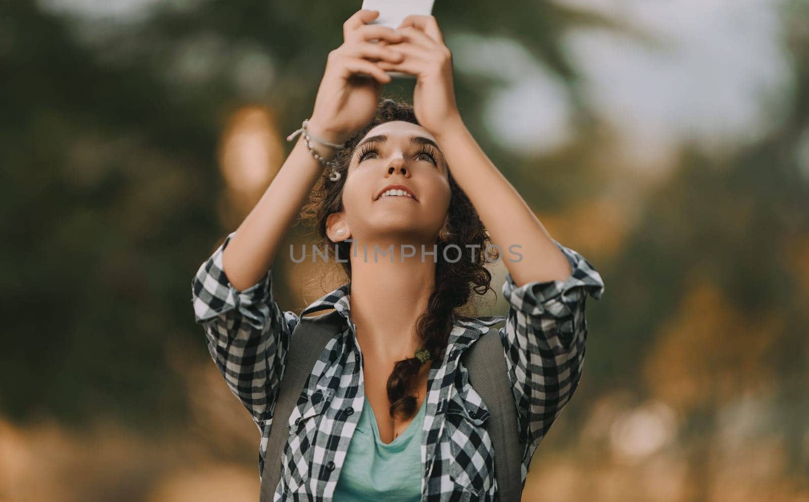 Woman with curly braided hair and a checkered shirt holds a smartphone while looking up and smiling. Her relaxed posture and backpack suggest she is enjoying capturing a moment outdoors