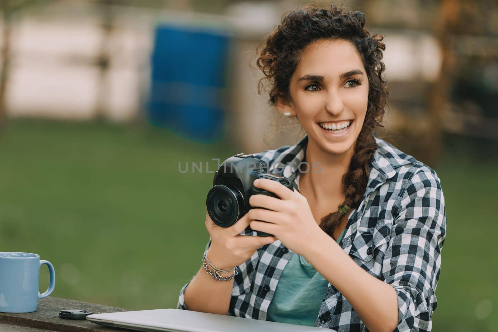 Young woman with curly hair in a braid, dressed in a checkered shirt, sits at a table outdoors, smiling while looking at her camera. Her laptop indicates a focused photography workflow
