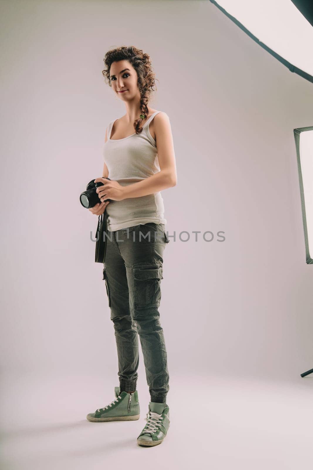 Confident woman with curly hair in a braid, wearing a sleeveless top and cargo pants, holds a camera. She stands in a studio, exuding a strong sense of pride and enthusiasm for her photography role