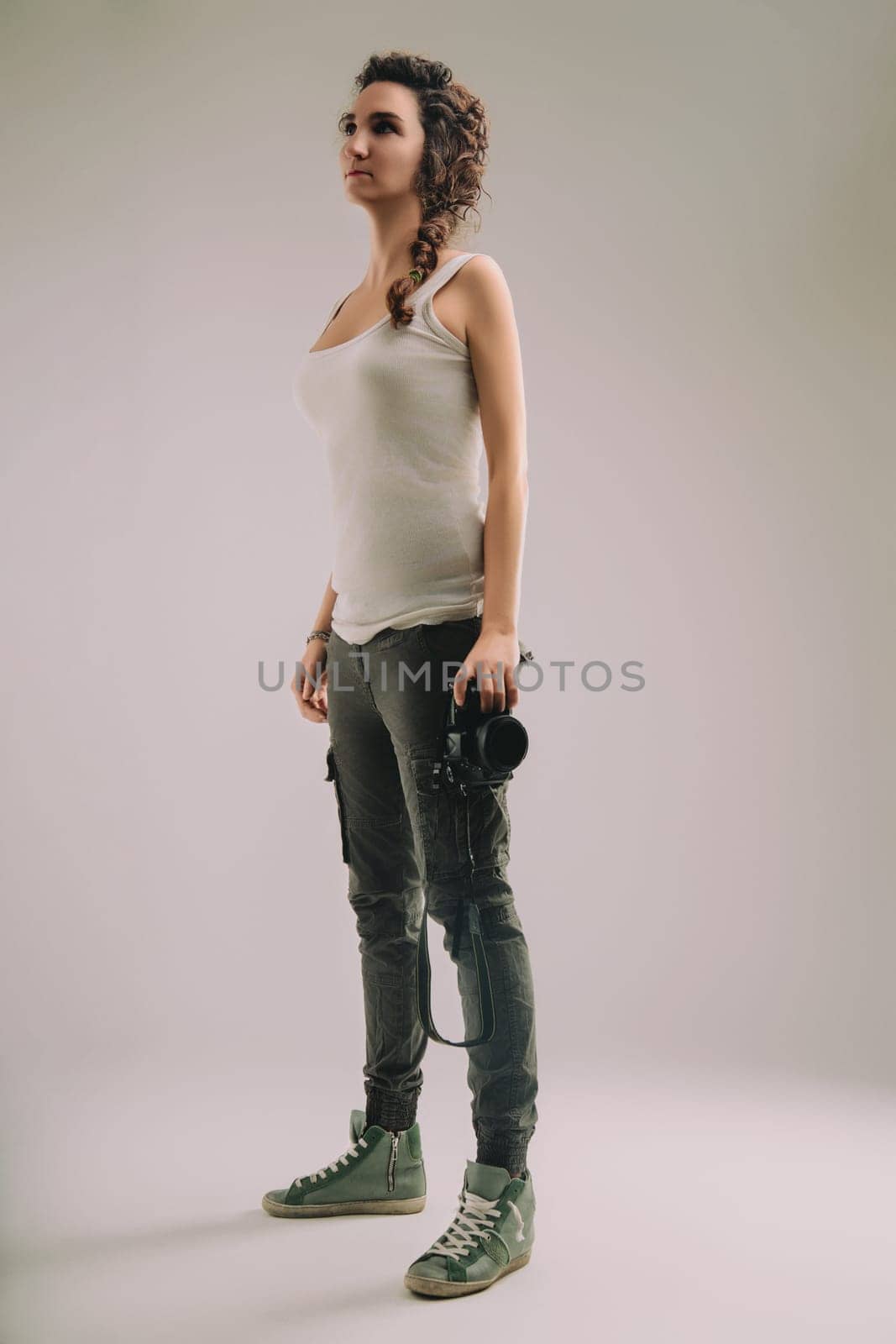 woman with curly braided hair and a sleeveless top, holding a camera, stands confidently in a studio. Her demeanor showcases her strong belief in the importance and coolness of her photographer role