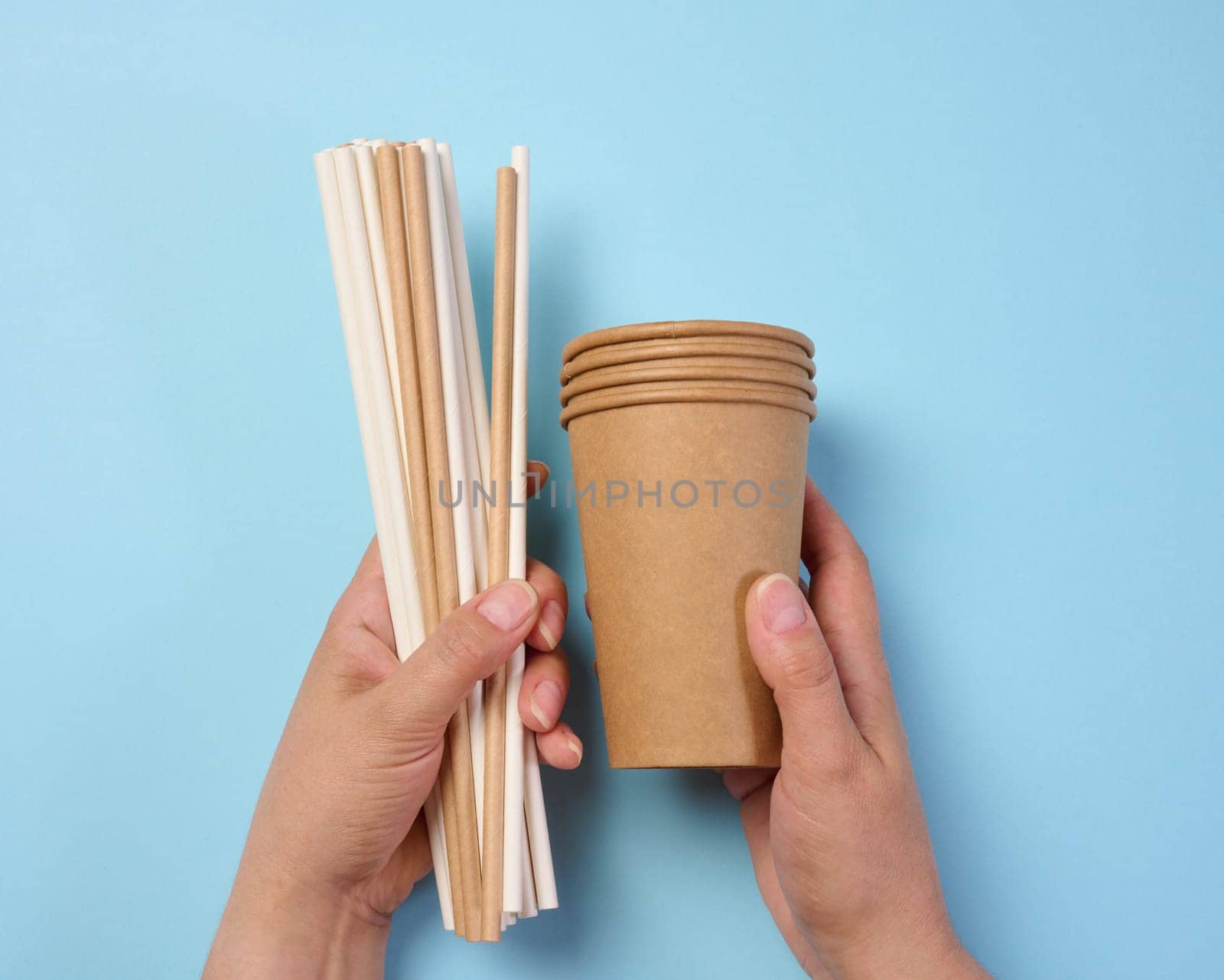 Female hand holding paper cocktail straws and paper cups on a blue background, top view