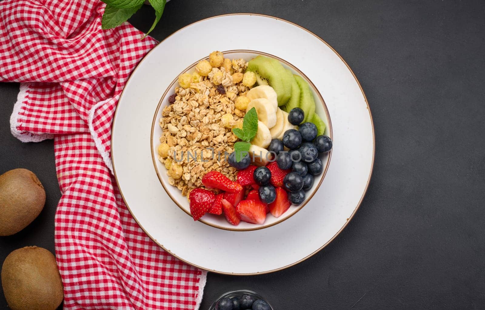Granola with strawberries, kiwi, banana and blueberries in a round plate on a black table. Healthy and tasty food, top view
