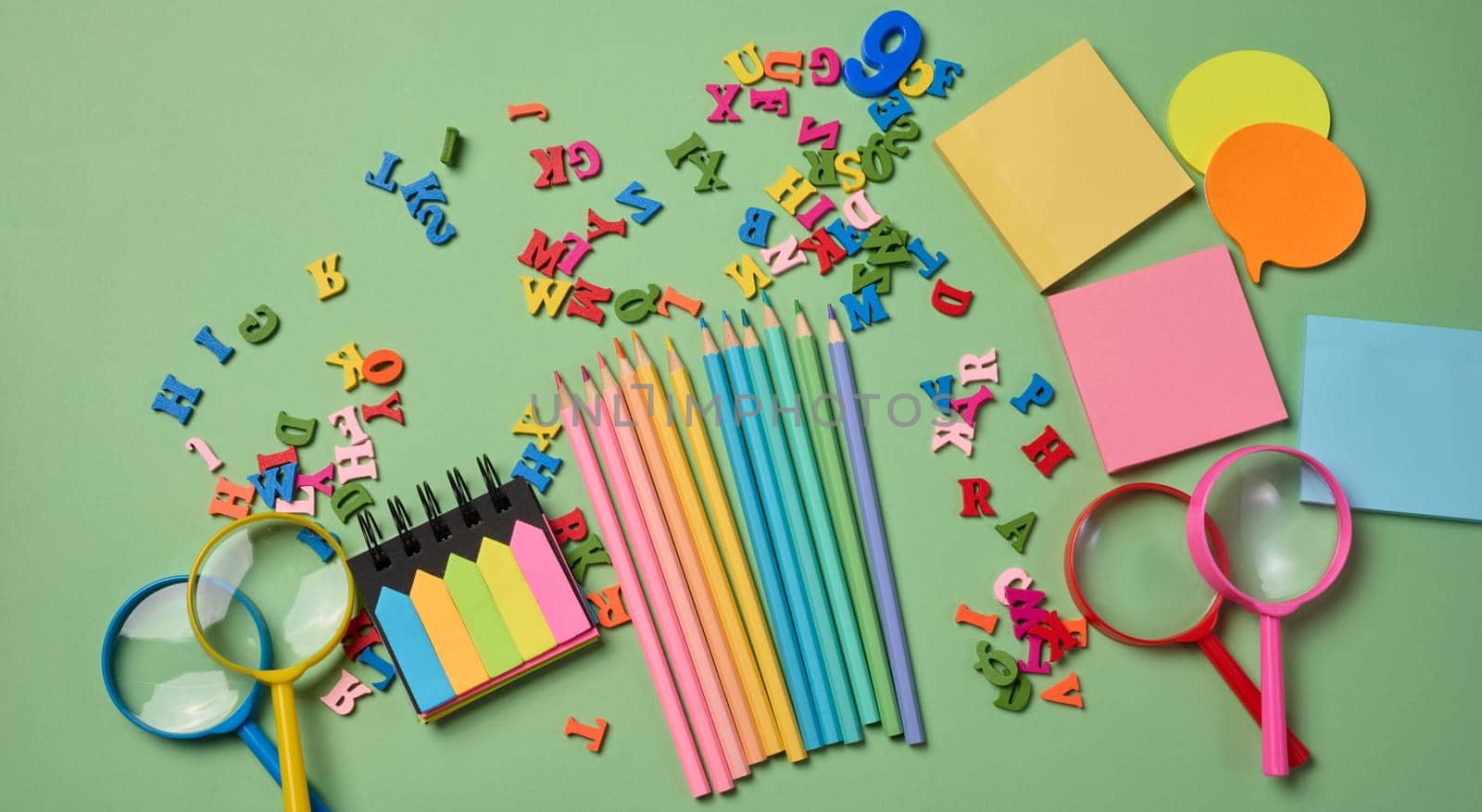 Stationery laid out on a green background. Multi-colored pencils ,  sticky notes and wooden letters, preparing for study or work, organizing workspace