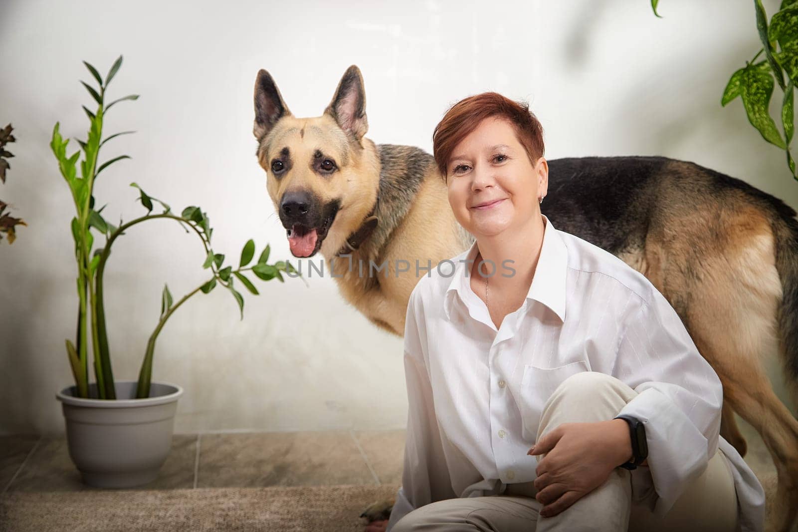 A middle-aged woman bonds with her German Shepherd dog in a comfortable, warm room filled with plants.