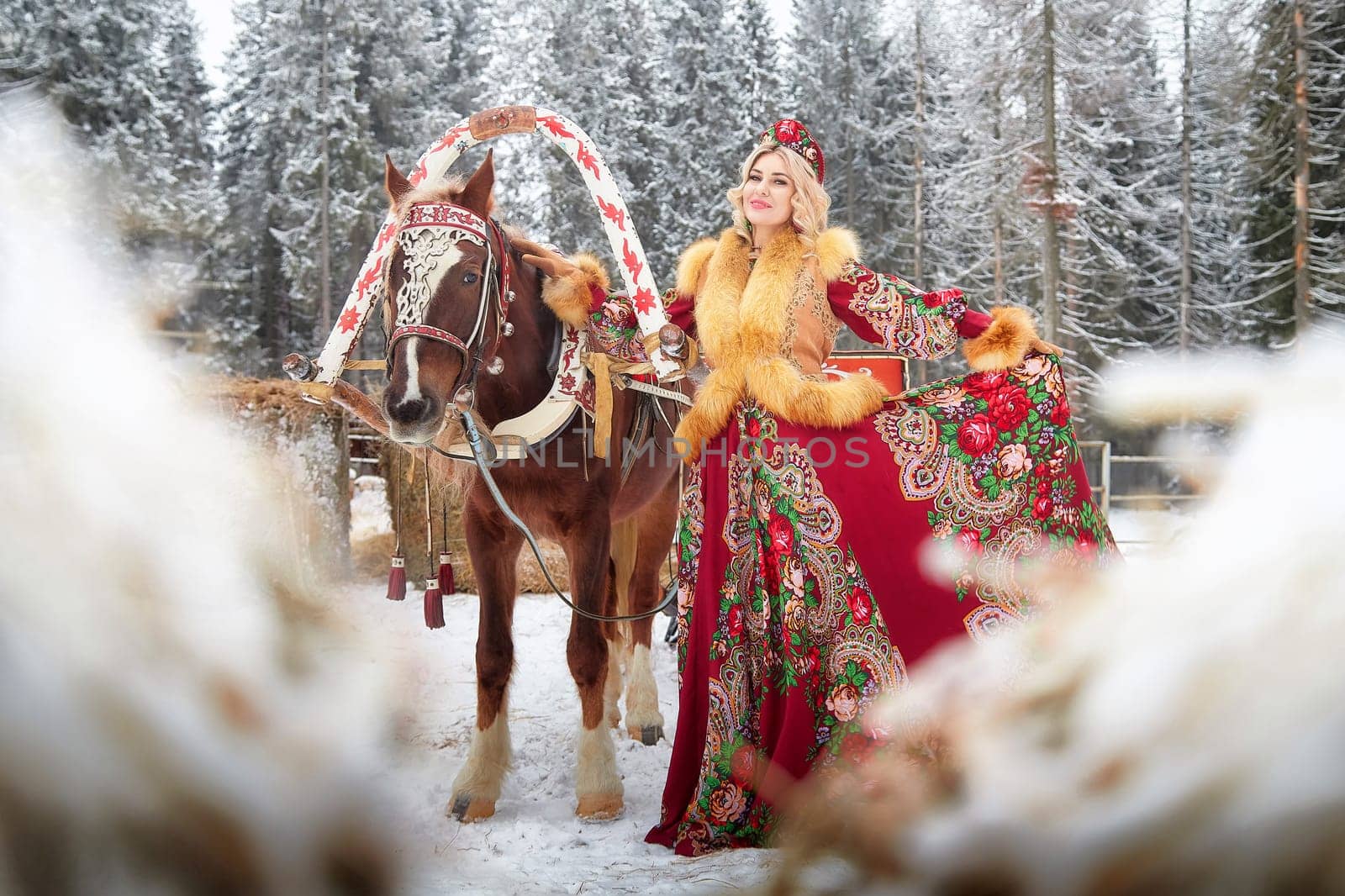 A stunning young woman in vibrant national attire interacts with a horse, celebrating Maslenitsa in a snowy setting.