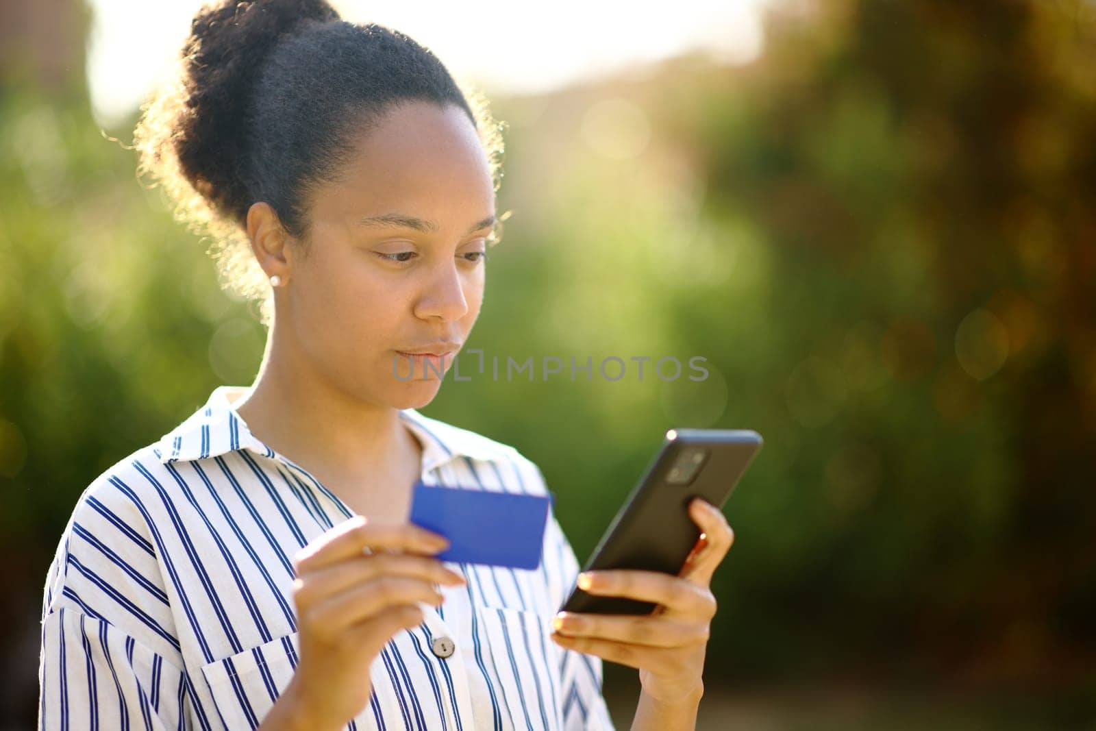 Serious black woman buying online using phone and credit card in a park