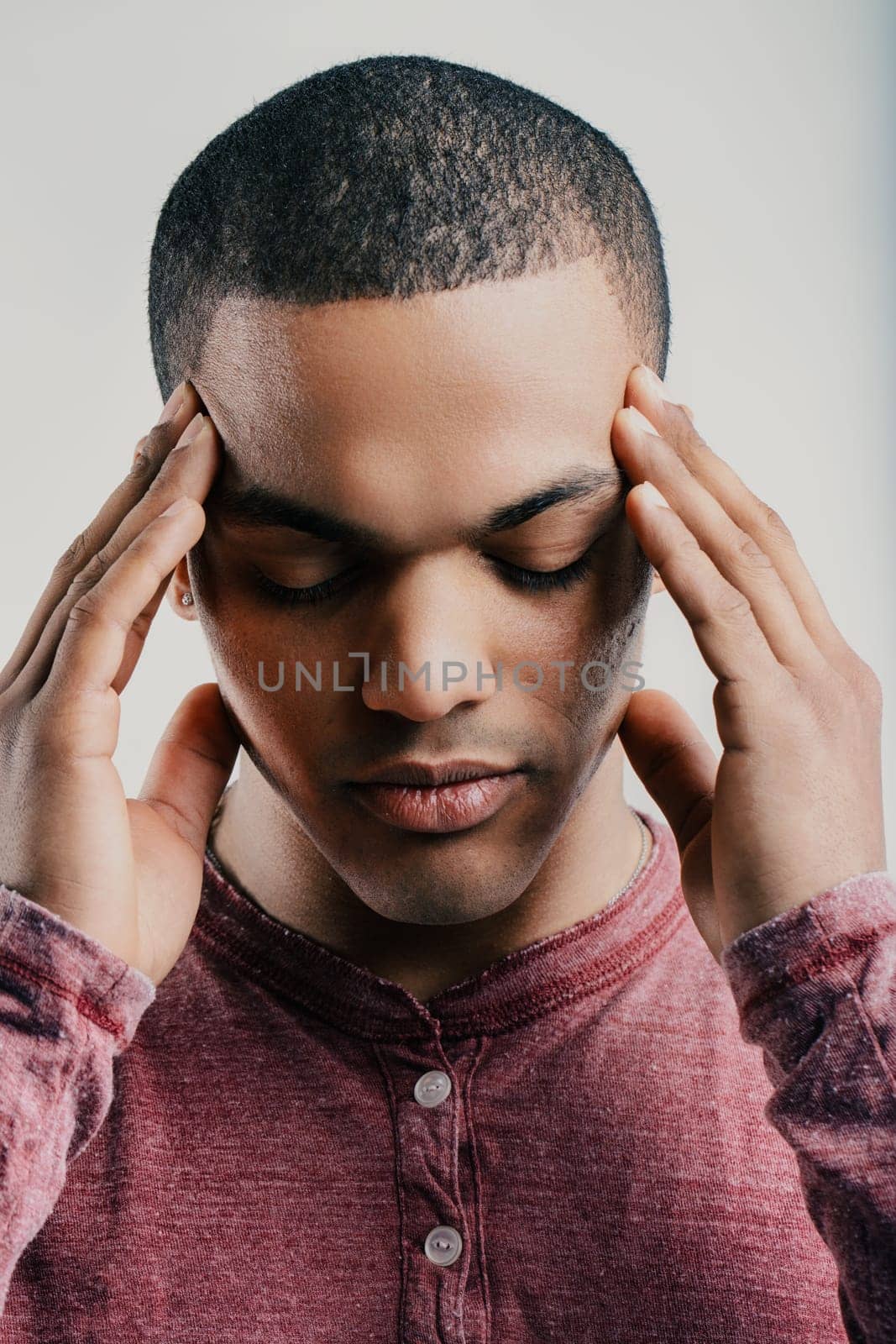 Young man with a pained expression, pressing his fingers to his temples. Wearing a maroon shirt, he appears to be experiencing a headache or intense stress, reflecting discomfort and frustration