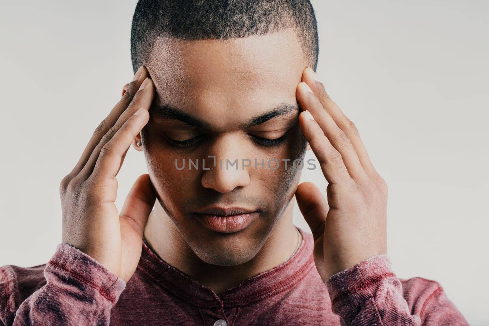 Pained young man, pressing his fingers to his temples while grimacing. Dressed in a maroon shirt, his expression suggests a headache or mental strain, indicating intense discomfort
