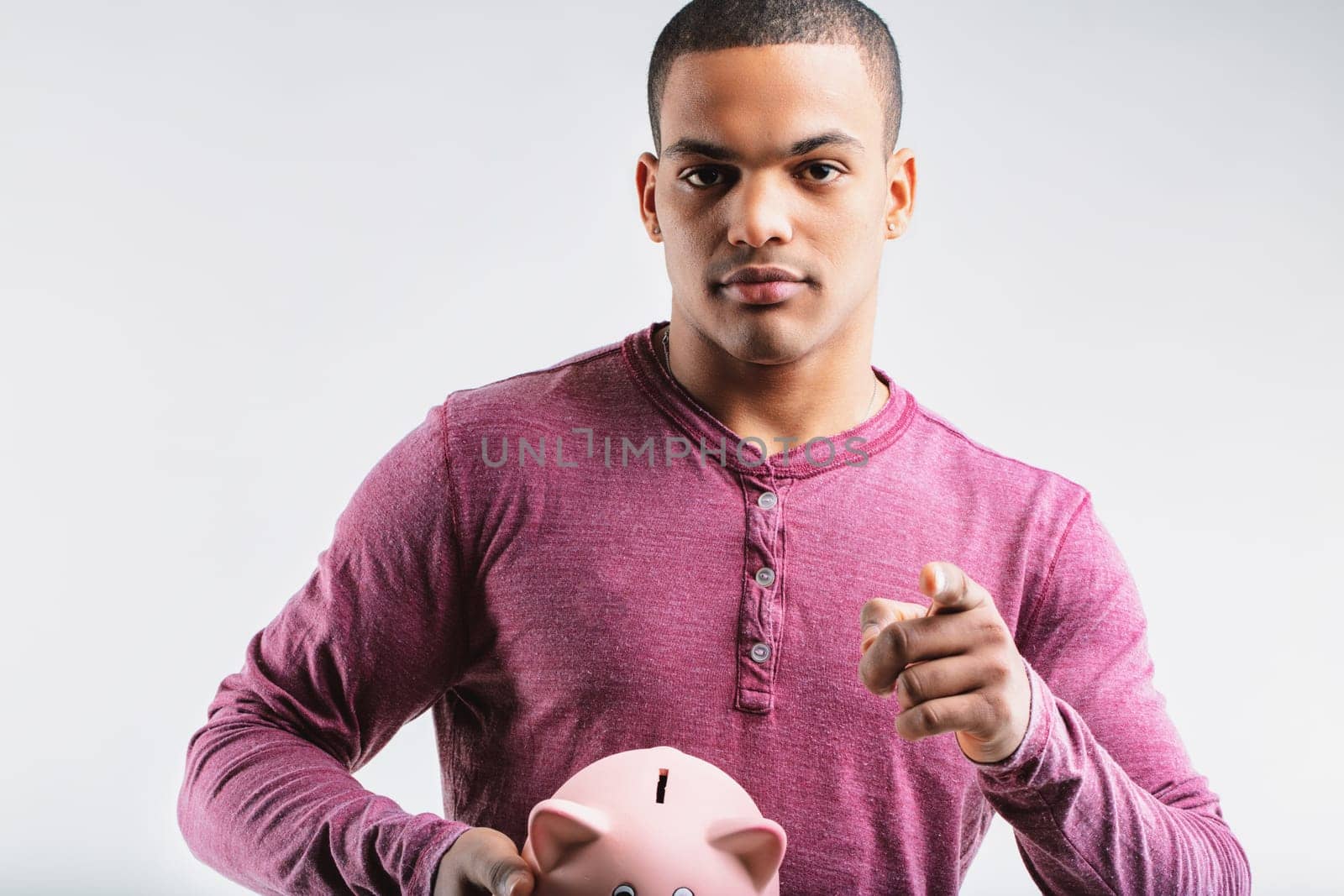 Young man in a maroon henley shirt, holding a piggy bank and pointing forward. His serious expression suggests advising caution with savings and the importance of investing wisely to avoid losses