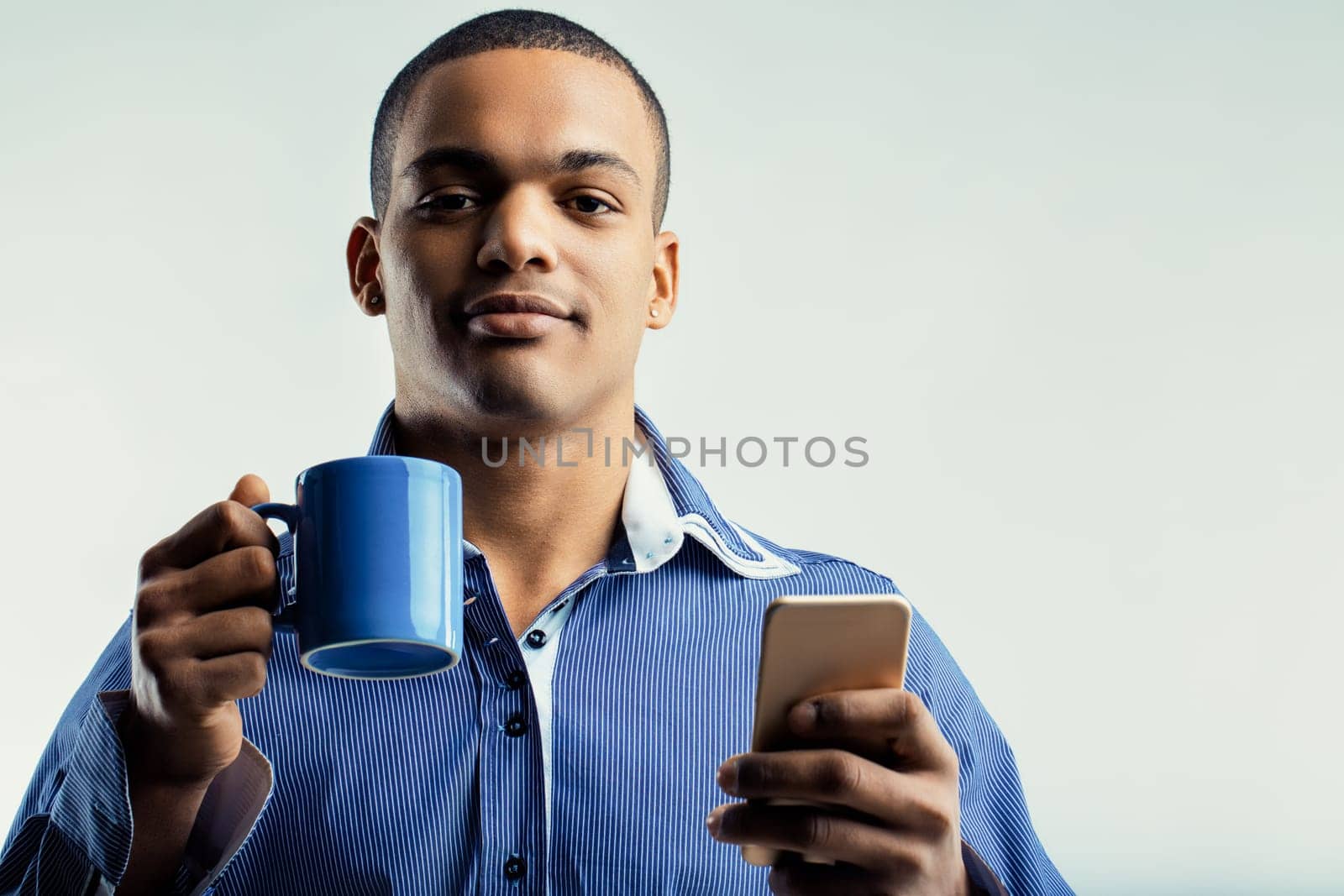 Man in a blue striped shirt, calmly holding a coffee mug and a smartphone. His relaxed demeanor suggests he is taking a break while staying connected through his phone