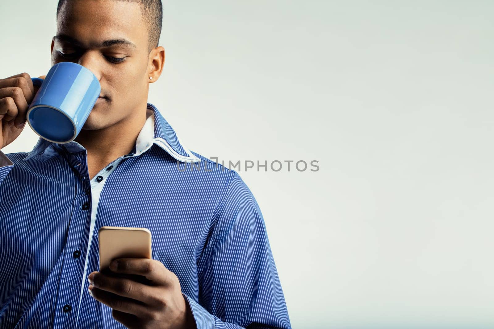 Individual with a serene look, holding a blue coffee mug and a smartphone. Dressed in a blue shirt, he seems to be enjoying a peaceful moment and staying informed