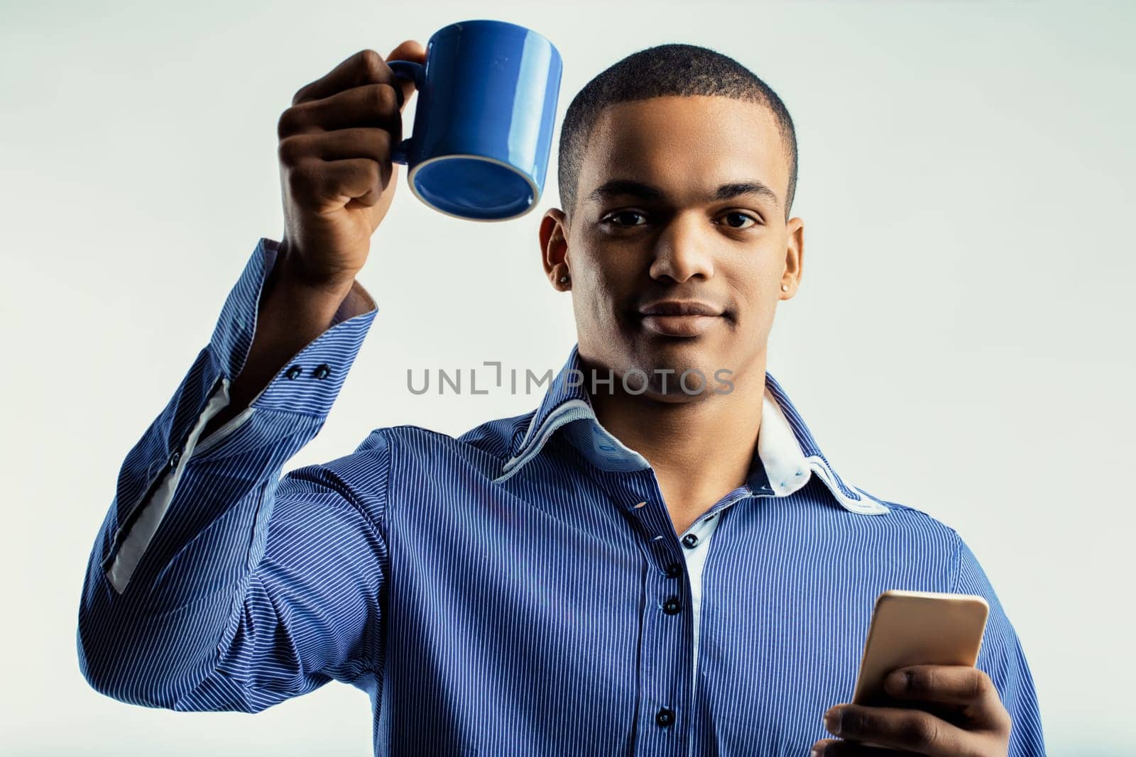 Calm young man, holding a blue coffee mug and a smartphone. His blue striped shirt and relaxed expression indicate he is enjoying a break while remaining connected