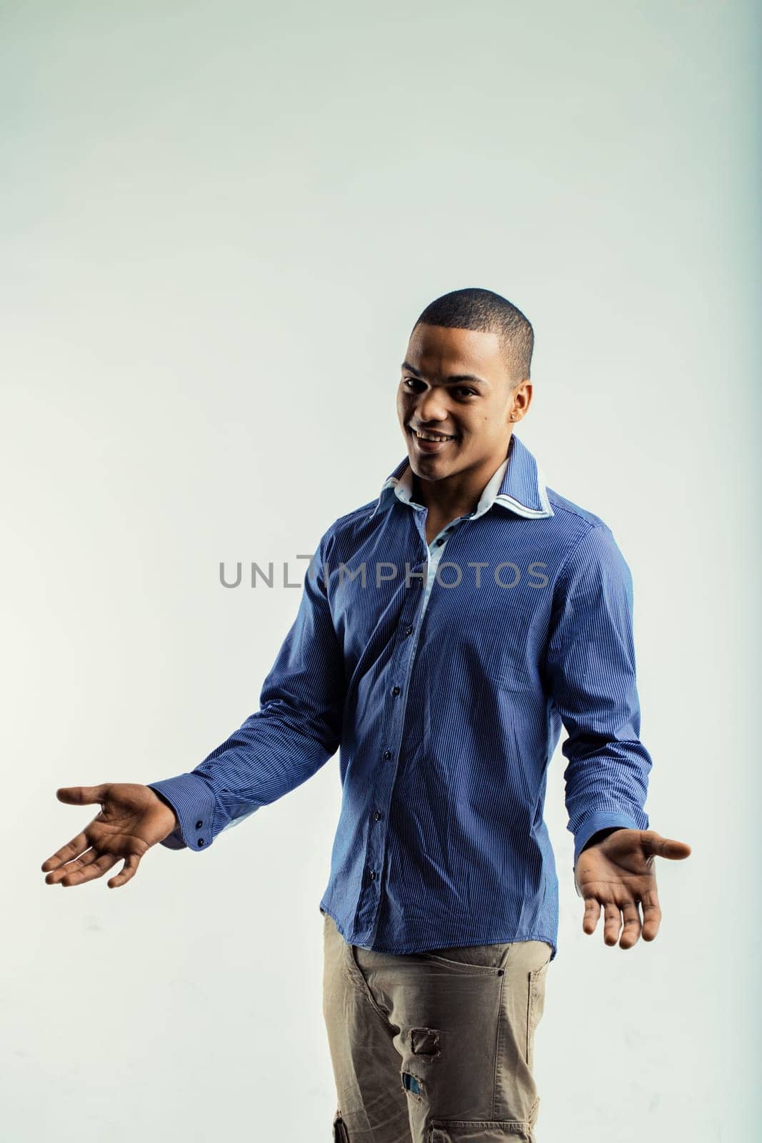 Young man in a blue striped shirt and beige pants, standing with arms open and palms up. His friendly expression and casual attire suggest he is inviting or welcoming, conveying openness