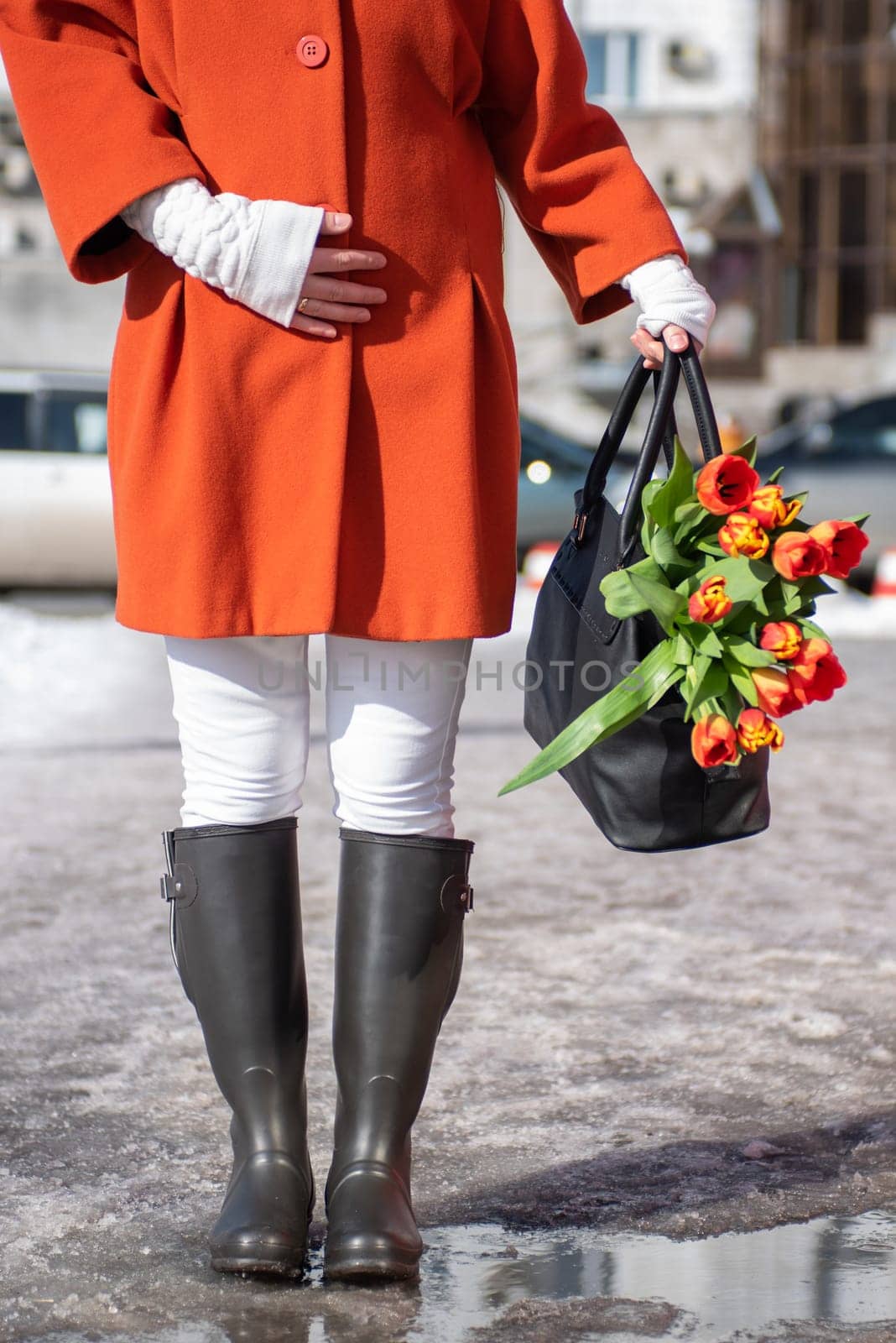 Closeup of woman in a orange coat and rubber boots walking down the street with a bouquet of tulips in spring city