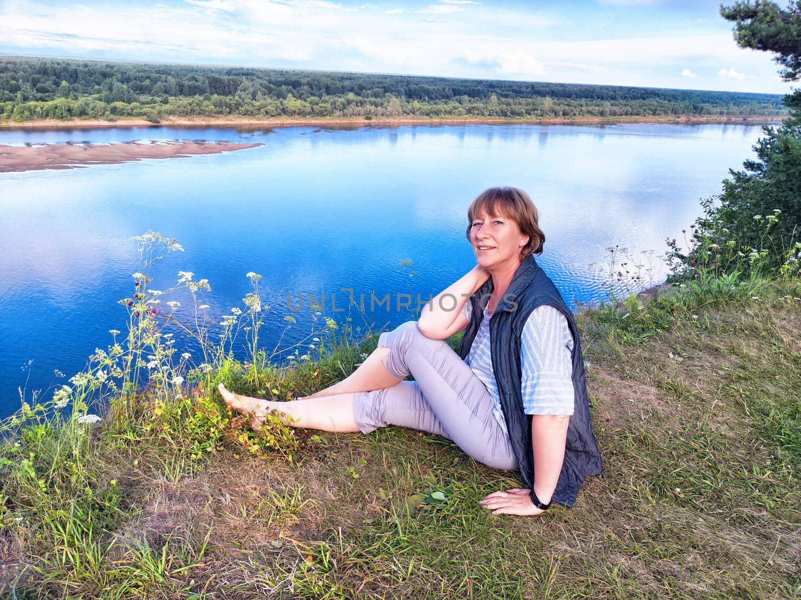 A middle-aged woman takes a selfie while relaxing by a tranquil river, surrounded by lush nature on a sunny day.