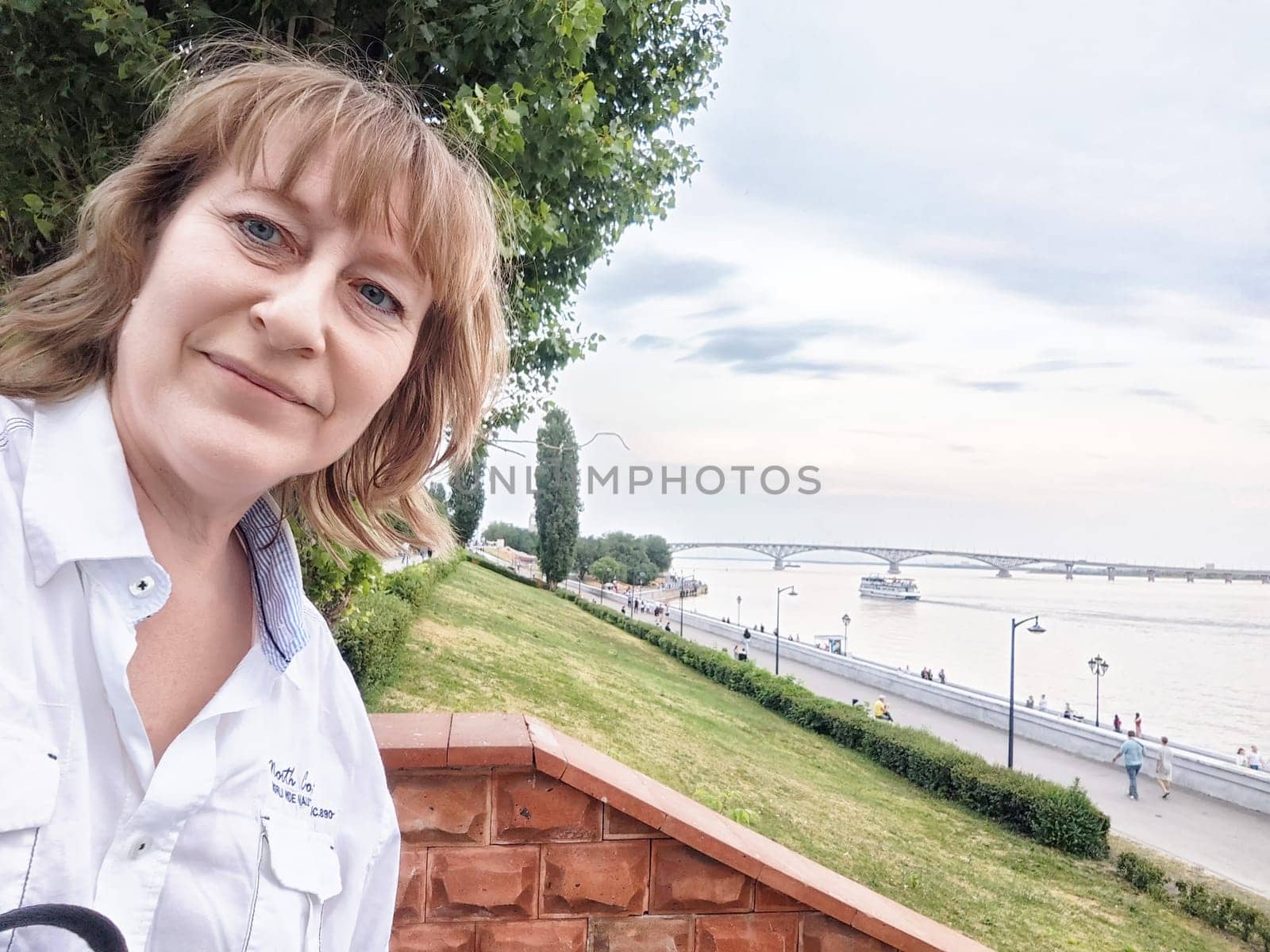 A mature blonde woman captures a cheerful selfie while exploring a picturesque riverfront area during sunset.