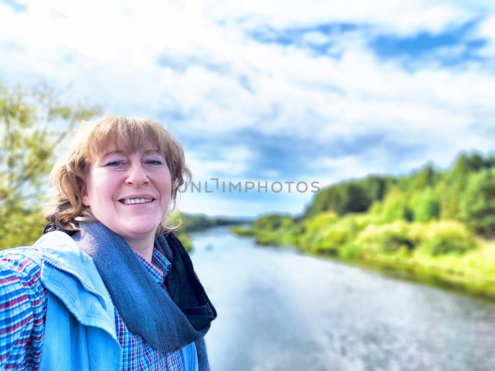 A middle-aged traveler blogger enjoys her excursion by the river, capturing a cheerful selfie amidst nature's beauty.