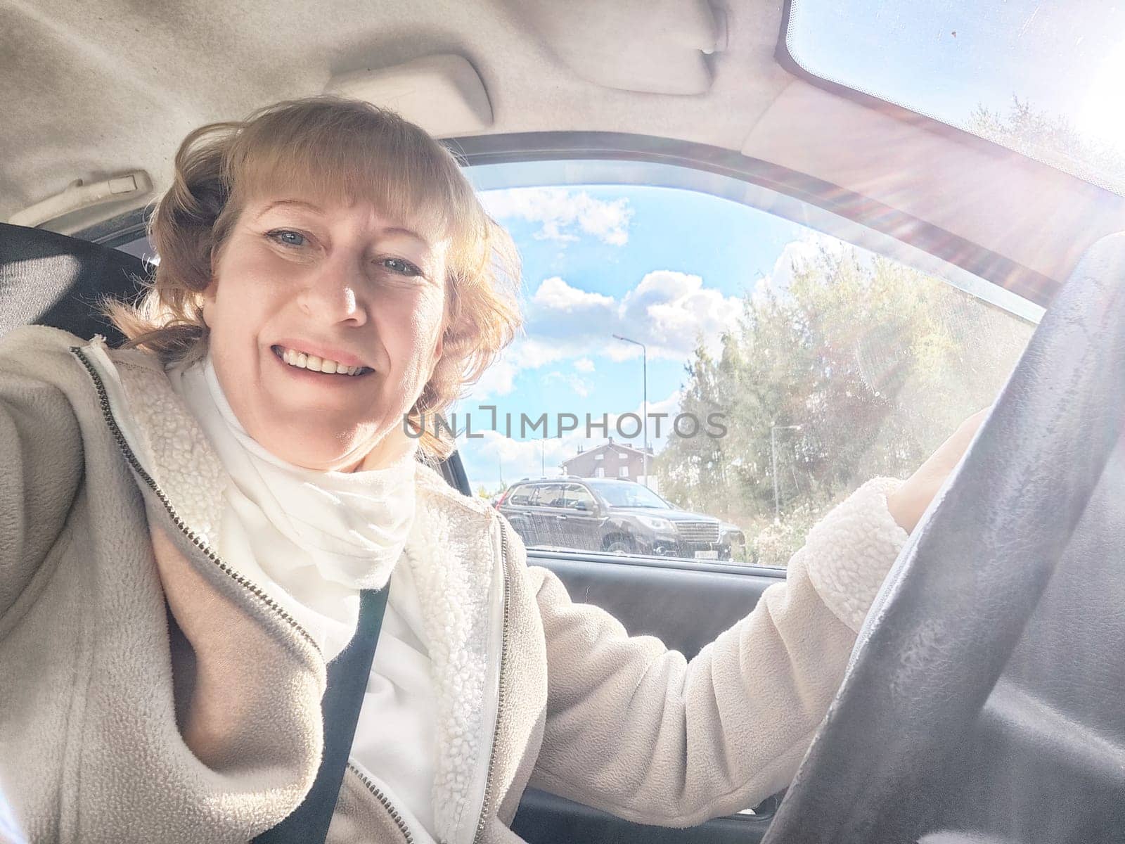 A middle-aged blonde woman smiles while driving, enjoying her adventure on a bright, sunny day.