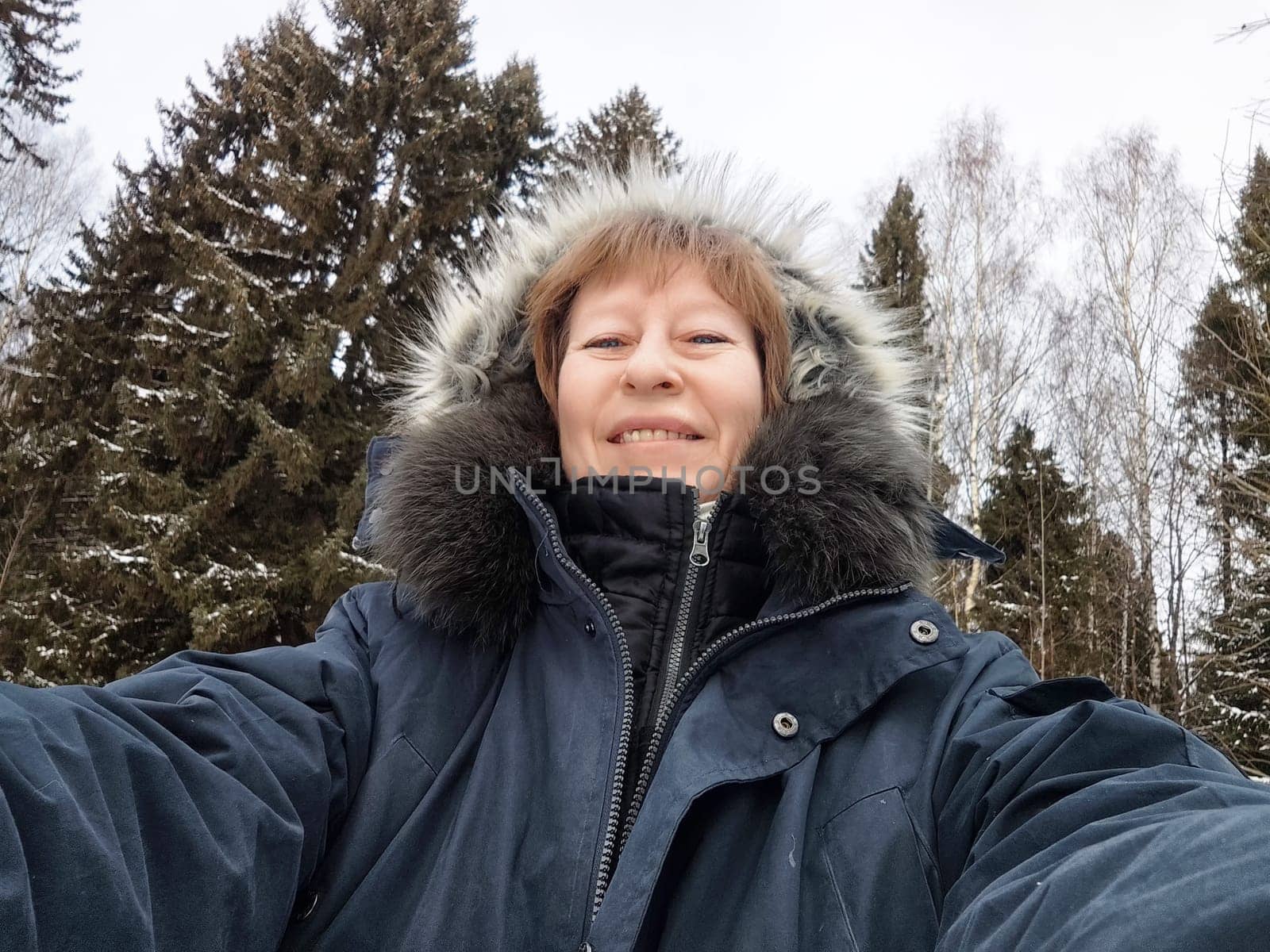 Blonde woman smiles while taking a selfie in a snowy forest, showcasing her vacation spirit and love for adventure.
