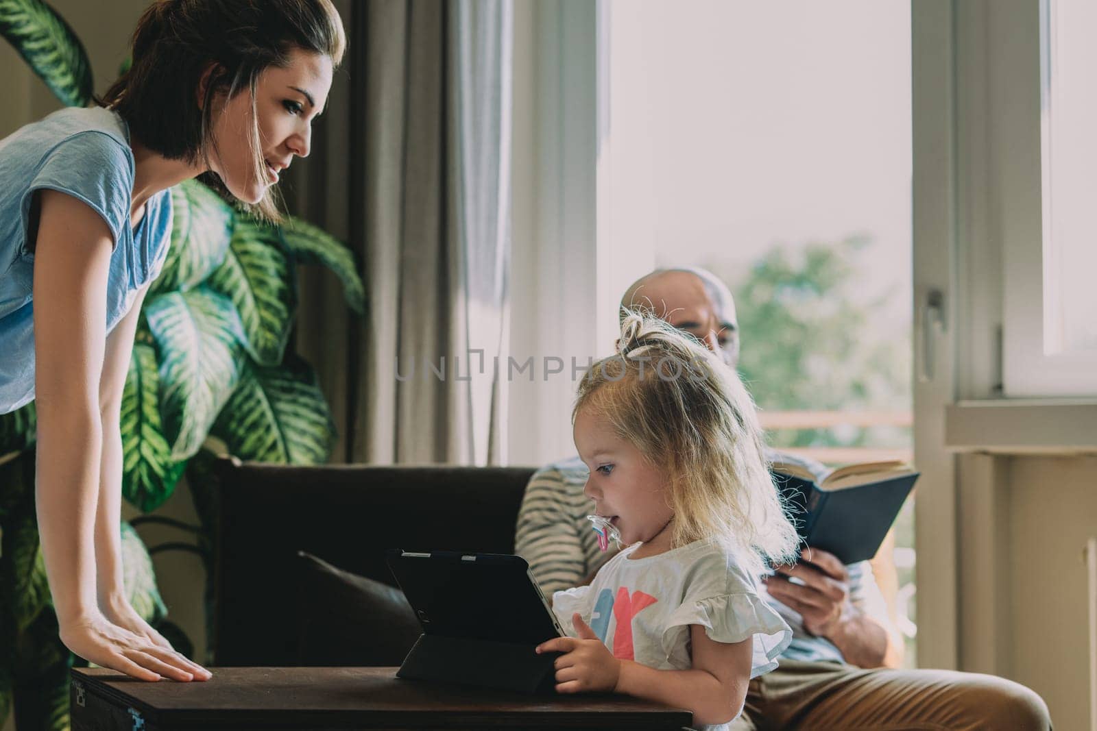 Young family is spending time together at home, with the mother watching over her daughter who is using a digital tablet while the father reads a book