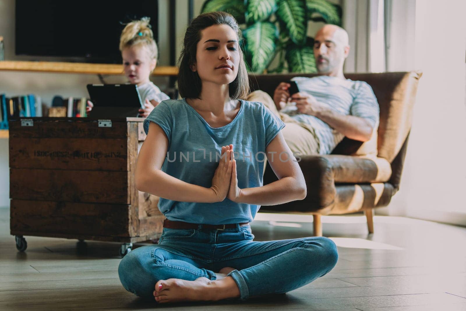 Young mother is maintaining her inner peace by practicing yoga and meditation in her living room while her family relaxes and uses technology