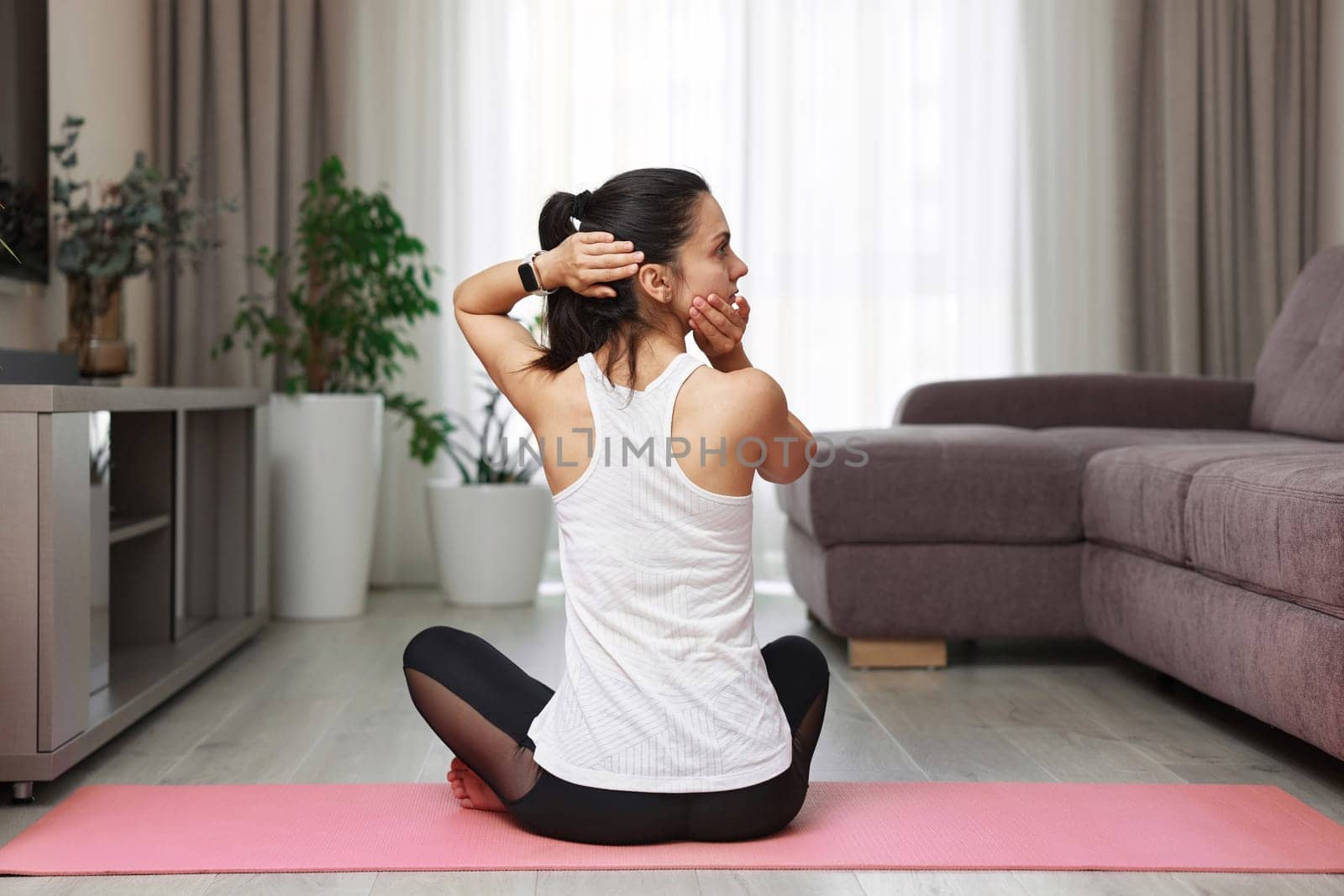 back view of Caucasian woman stretching head and warming up neck before workout sitting on yoga mat at home