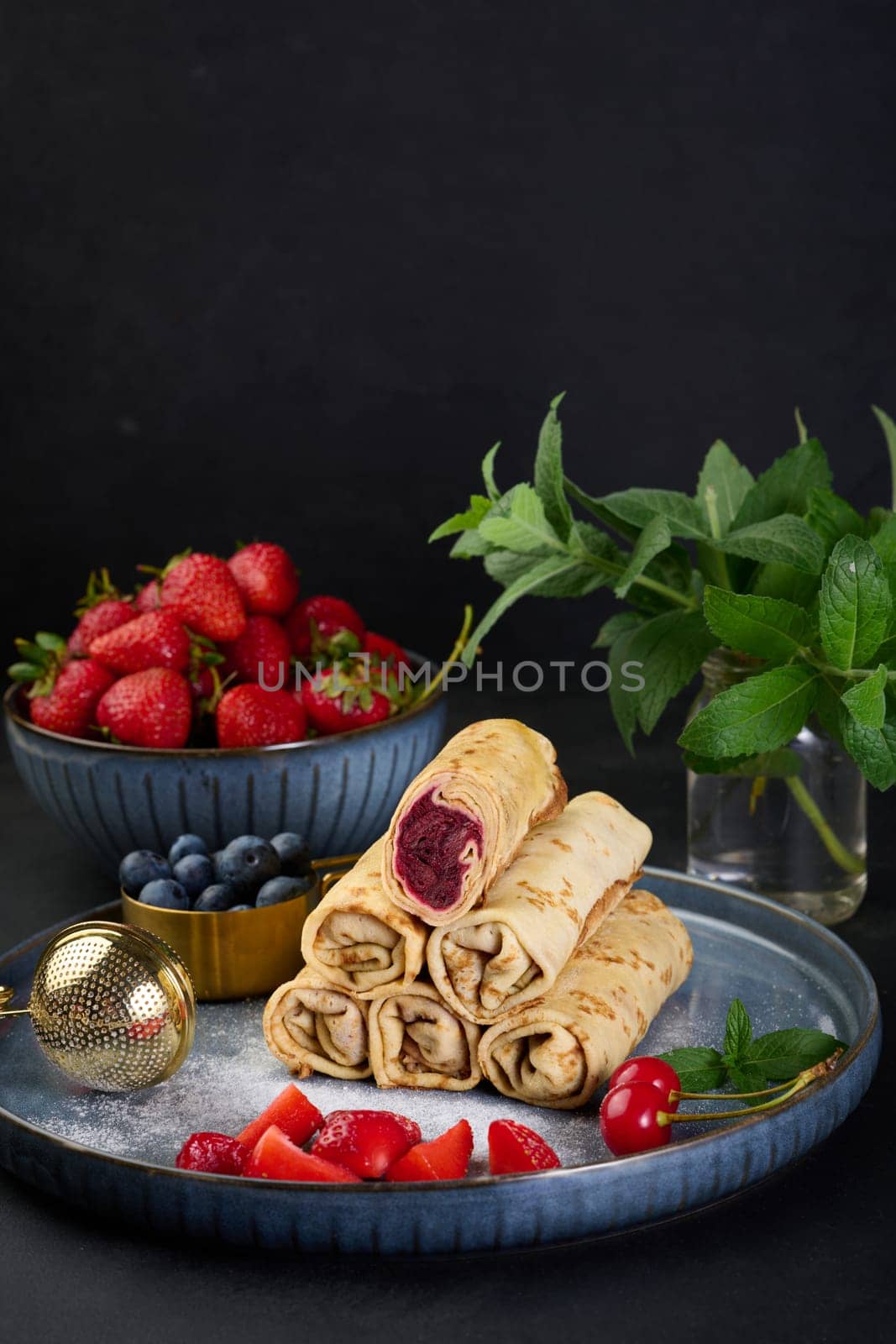 Crepes stacked in a pile, one of which is cut and filled with berry filling. The plate is also decorated with blueberries, strawberries, and cherries