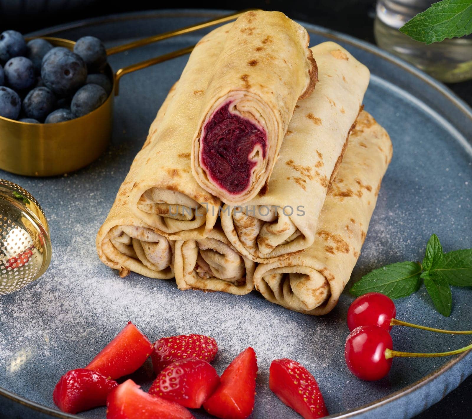 Crepes stacked in a pile, one of which is cut and filled with berry filling. The plate is also decorated with blueberries, strawberries, and cherries