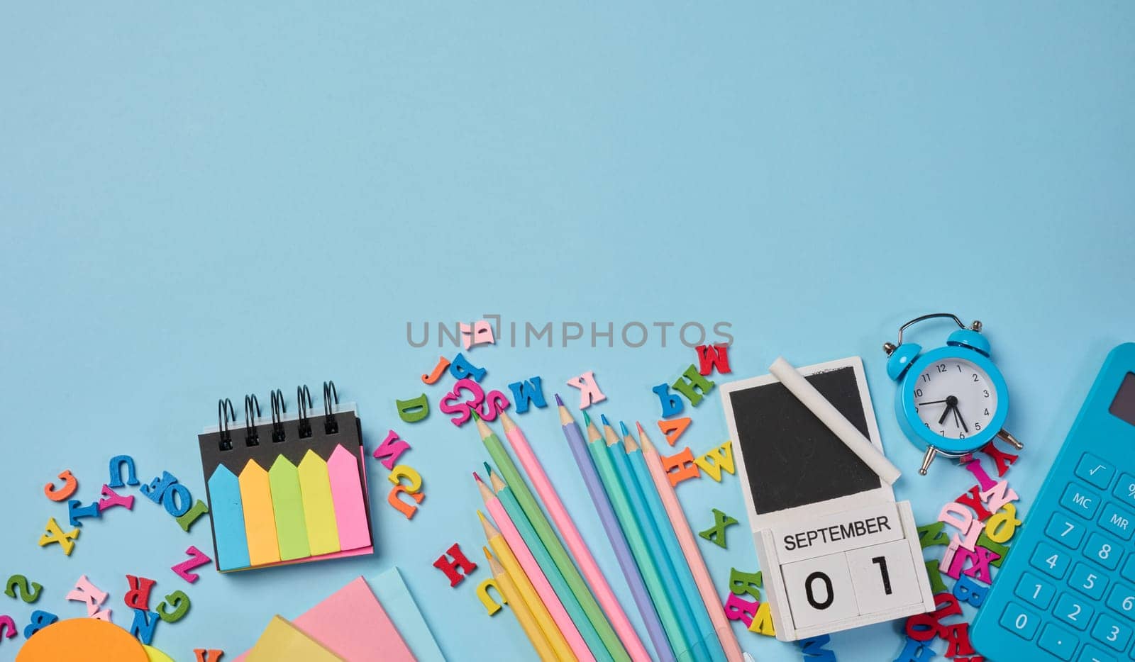 Stationery laid out on a blue background. Multi-colored felt-tip pens, calculator, alarm clock, sticky notes and wooden letters, preparing for study or work, organizing workspace