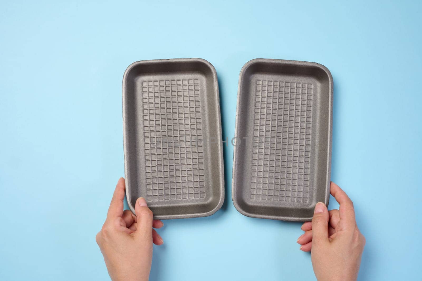 Hand holds black disposable polystyrene foam rectangular plate for packaging vegetables and meat on a blue background, top view