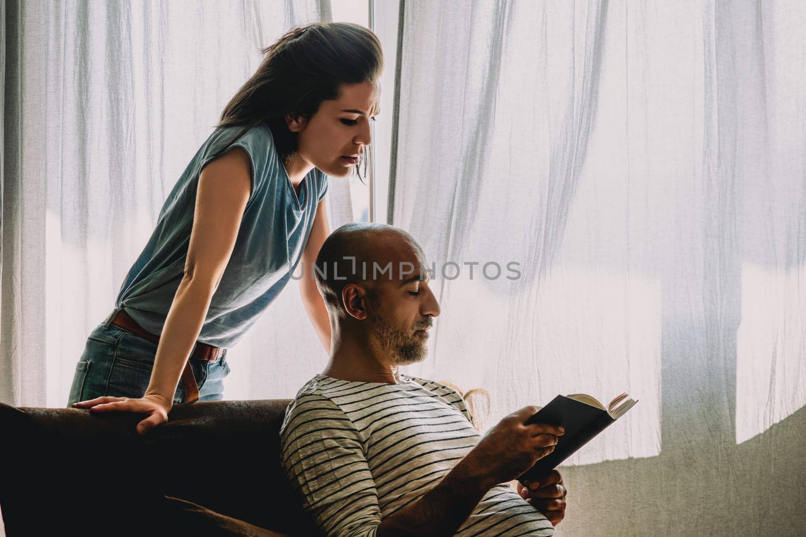 Man trying to avoid a confrontation with his angry wife by concentrating on reading a book, while she is trying to get his attention