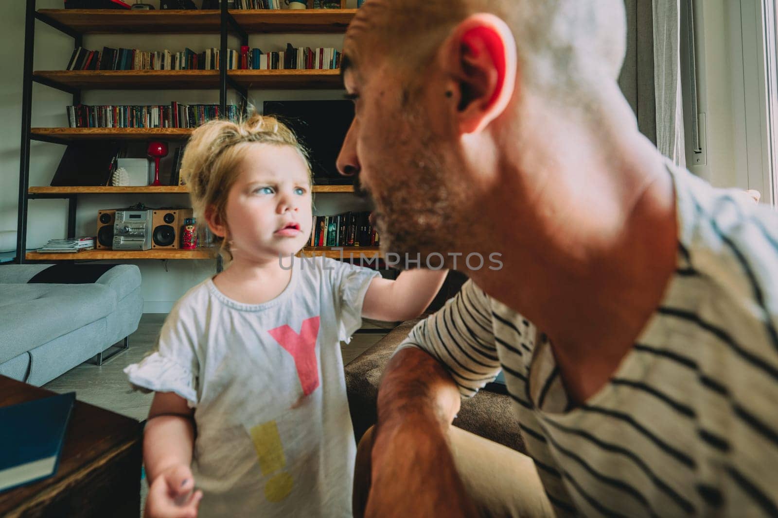 Little girl looking at her father while touching his face and asking him something in a living room with a bookcase in the background