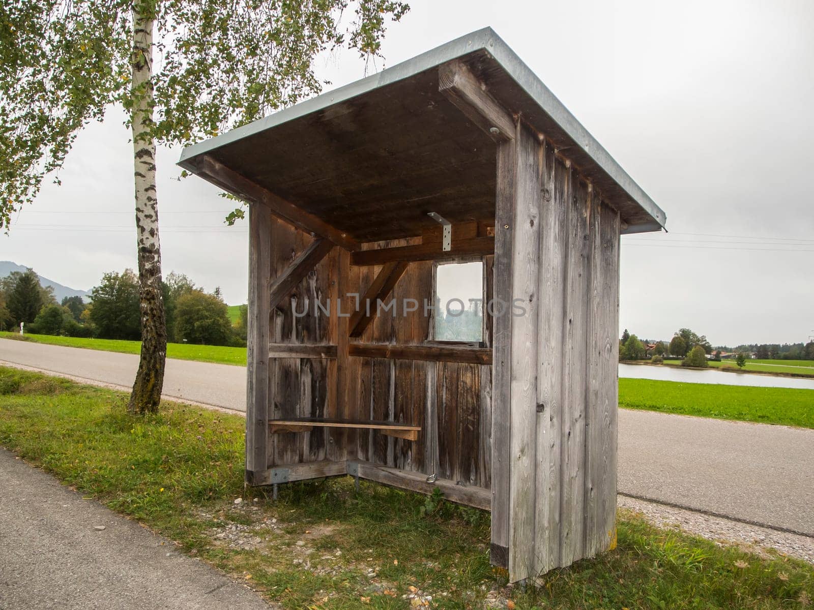 Landscape view with wooden bus shelter with adjacent birch tree under overcast sky in Bavaria.